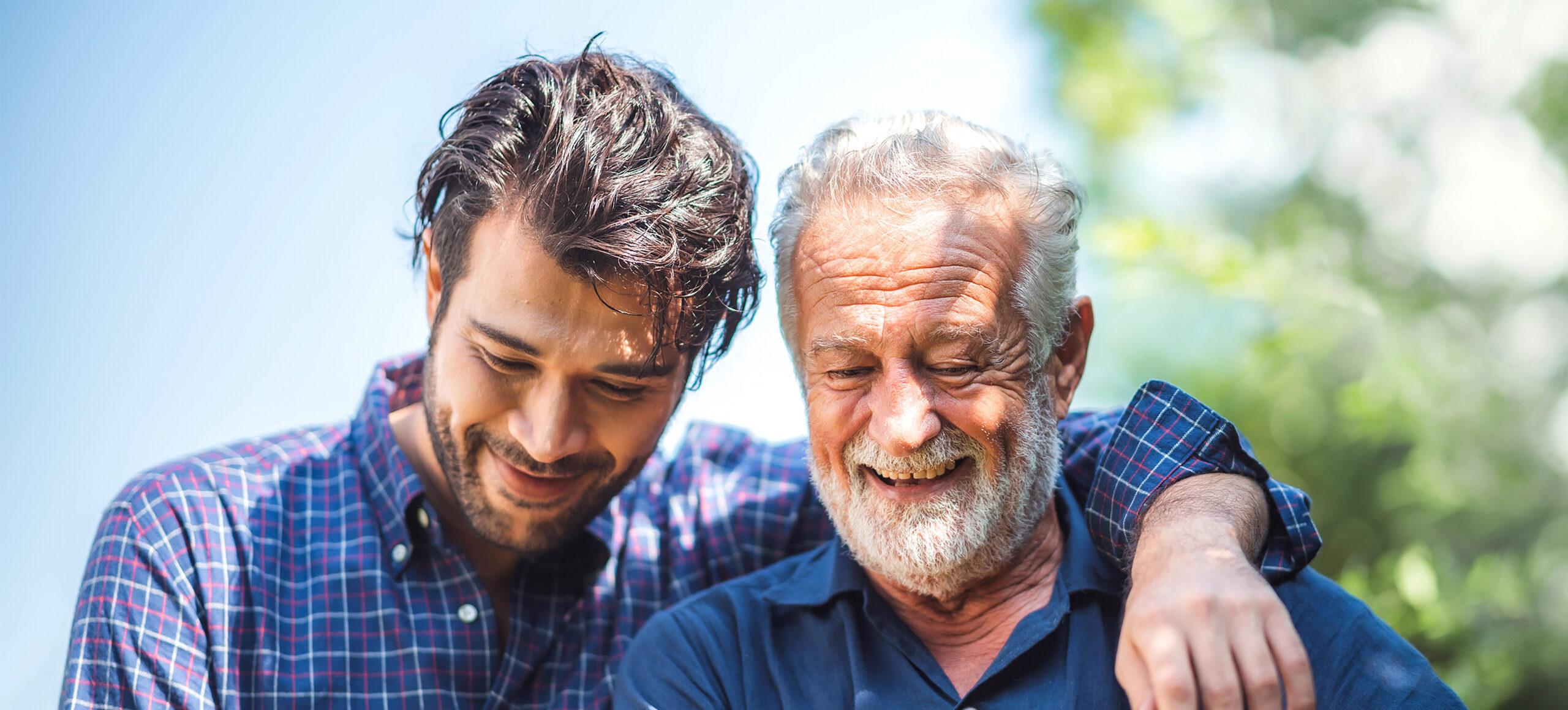 Adult son smiles with his arm around his father outdoors